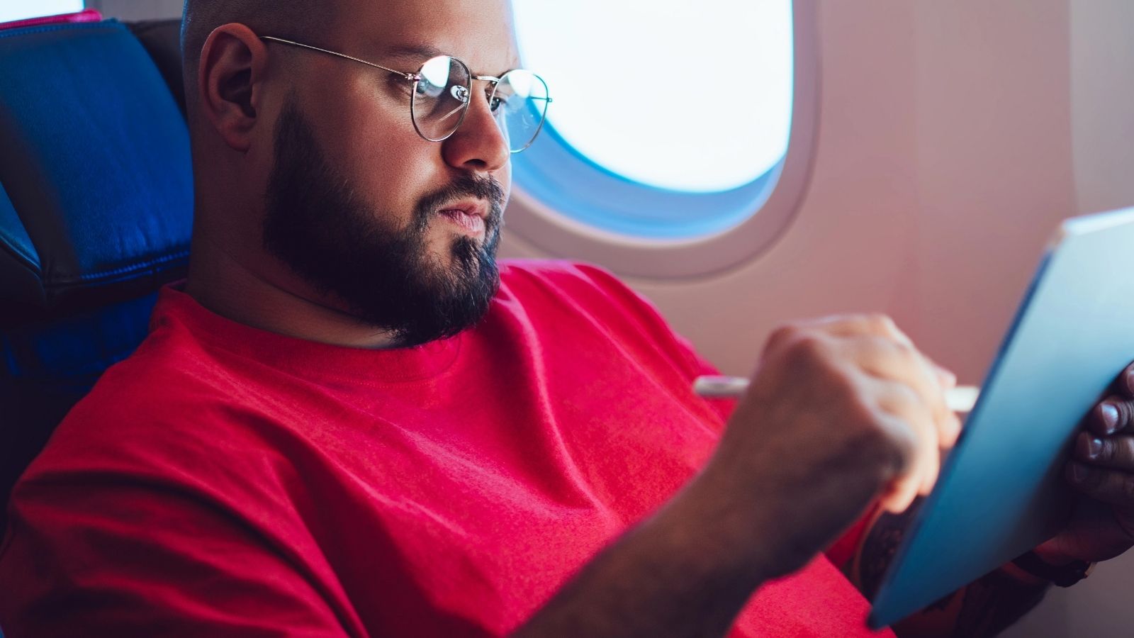 A man in glasses and a red shirt uses a stylus on a tablet beside an airplane window.