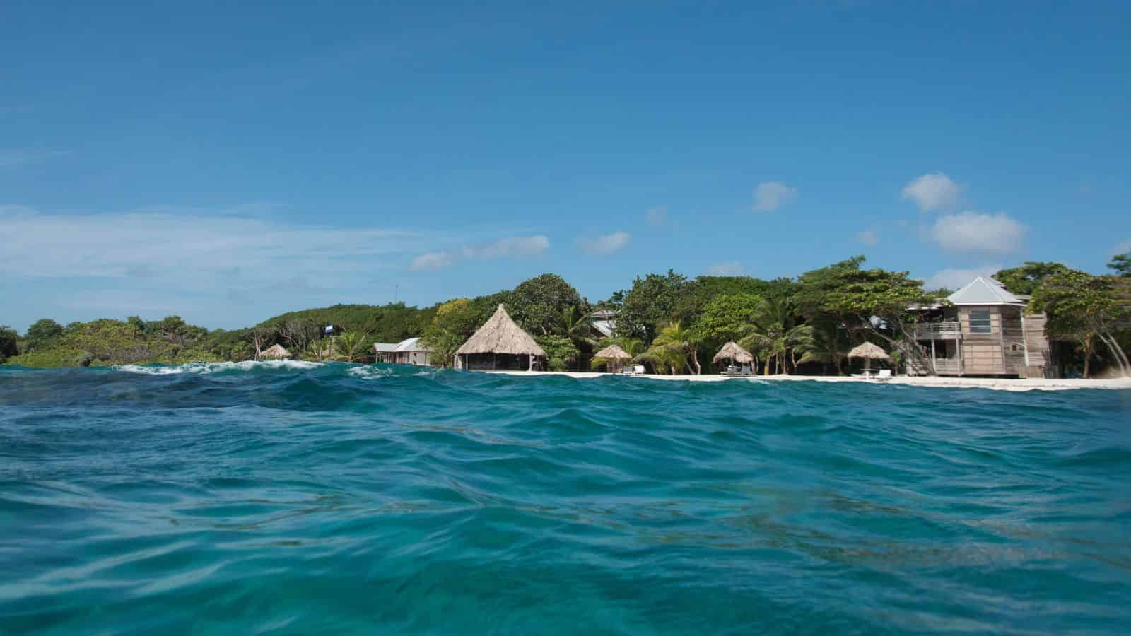 Clear blue water in the foreground with beach huts and palm trees along a tropical shoreline under a blue sky.
