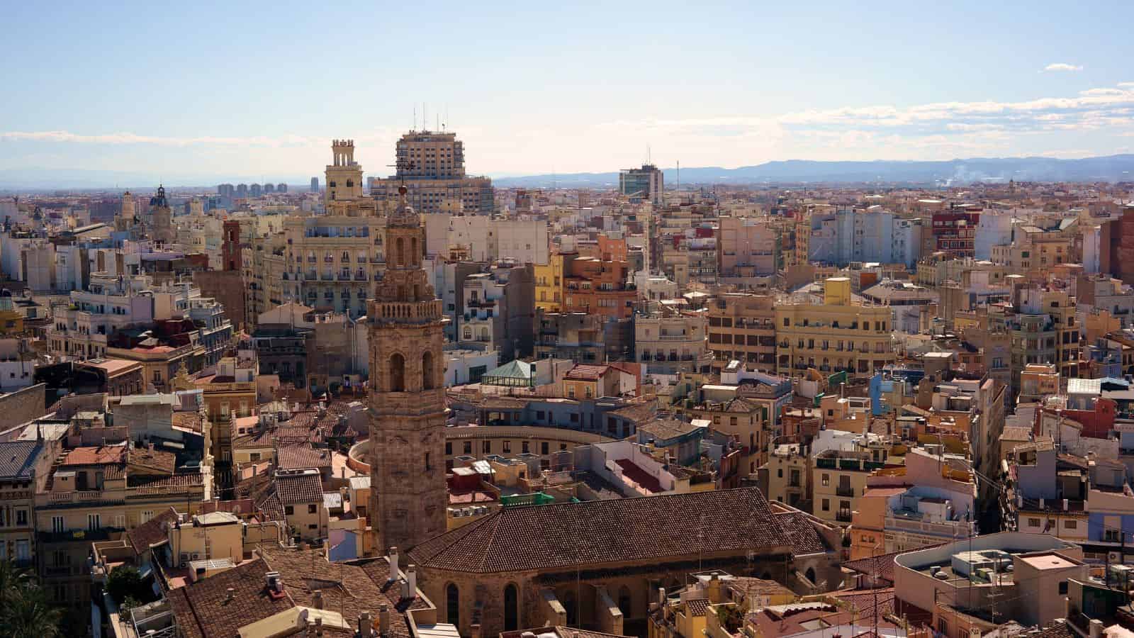 Aerial view of a dense city with a tall bell tower and colorful buildings beneath a clear sky.