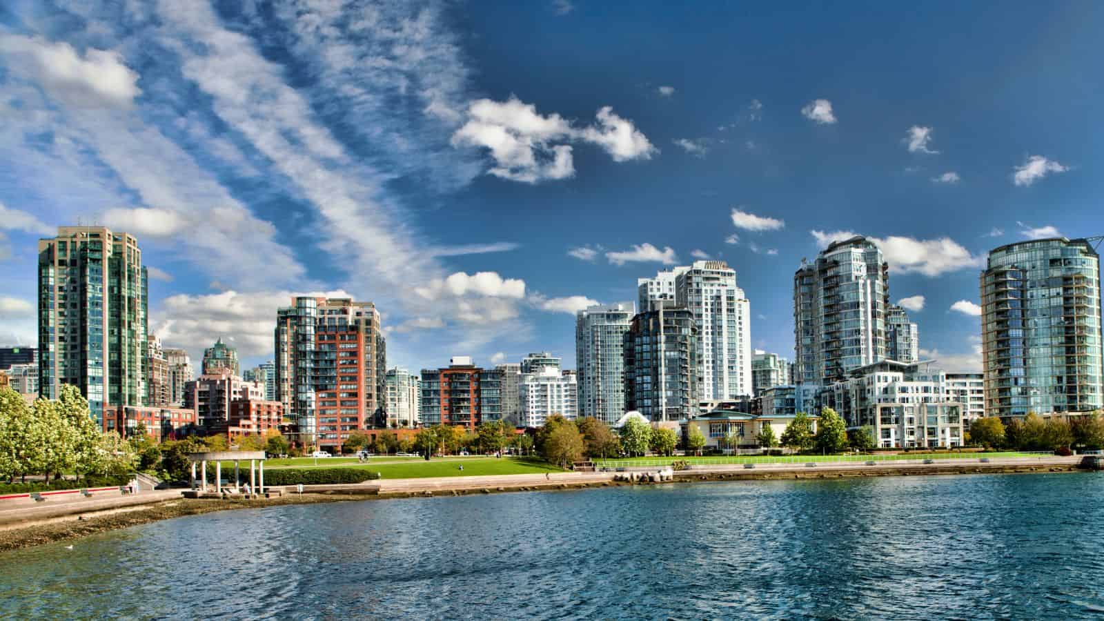 A city skyline of high-rises stands by a waterfront, with greenery on the shore and a blue sky dotted with clouds above.