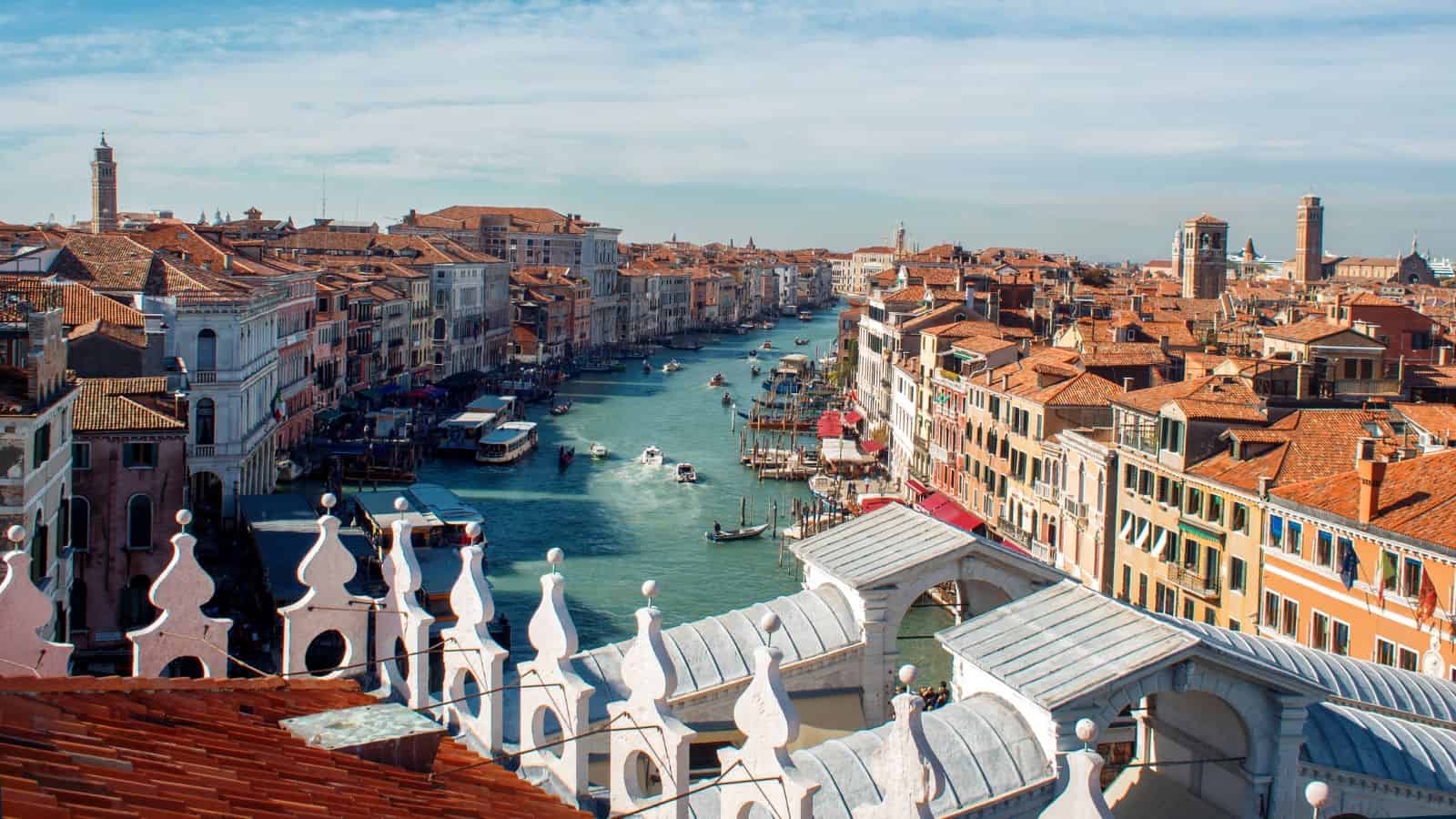 Aerial view of Venice’s Grand Canal lined with historic buildings and boats traveling on the water. Decorative white rooftop railings and red-tiled roofs are visible in the foreground.