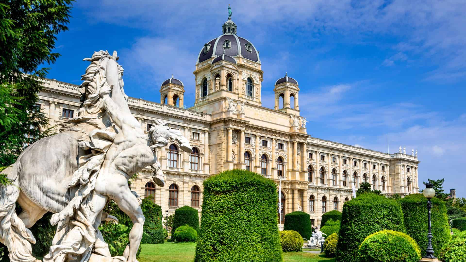 A grand domed historic building with ornate facade, trimmed bushes, and a rearing horse statue beneath a clear blue sky.