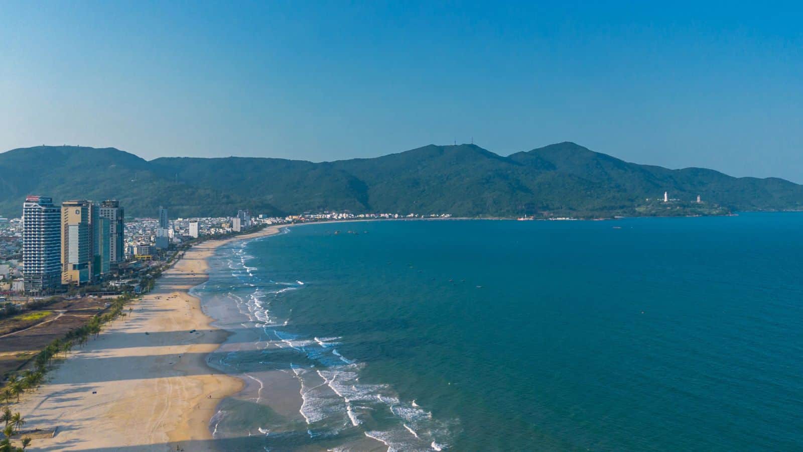 A wide sandy beach curves along the coastline with gentle waves, bordered by tall buildings on the left and green mountains in the background under a clear blue sky.