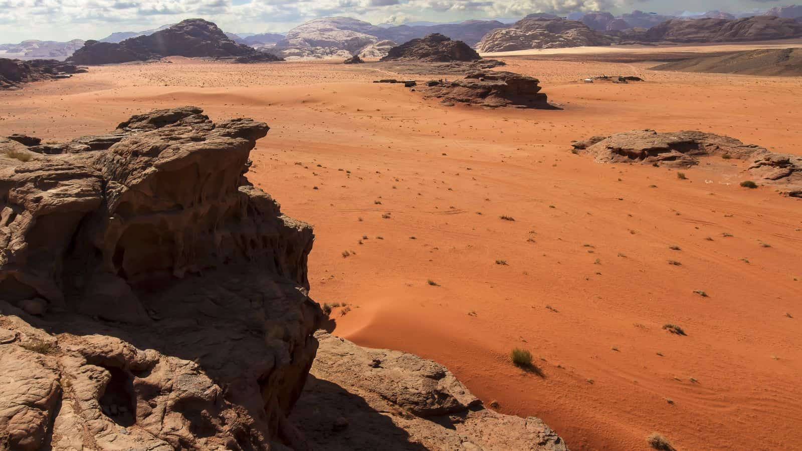 Rocky desert with red sand, scattered rocks, and distant mountains beneath a partly cloudy sky.
