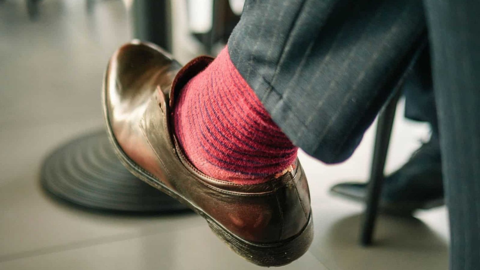 A person in shiny brown dress shoes, red striped socks, and dark pants sits with legs crossed.