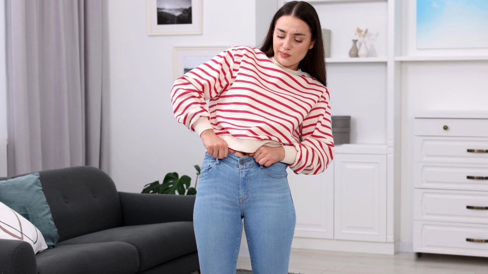 Woman in a red and white striped sweater struggles to button blue jeans in a living room.