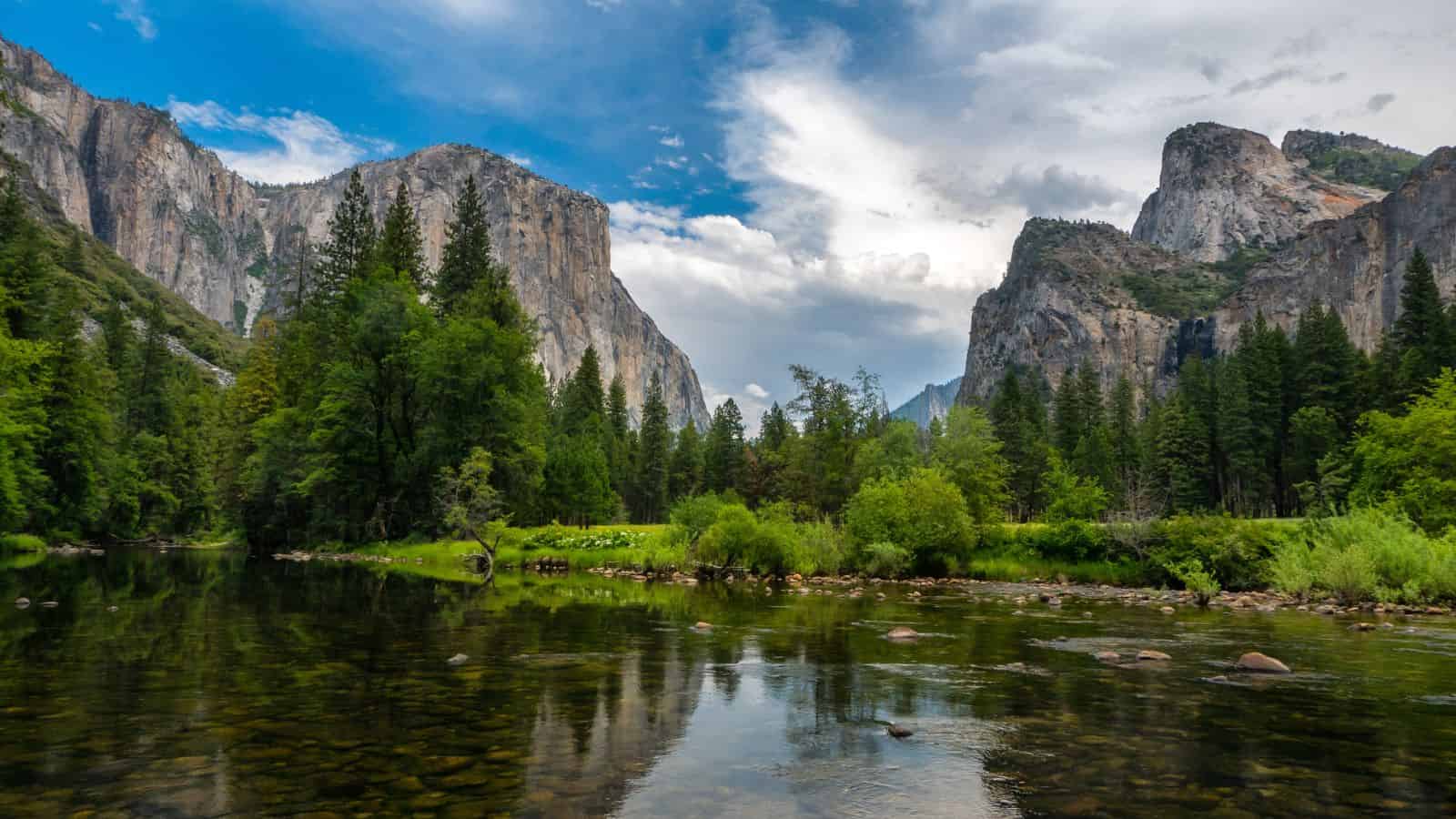 A serene river reflects the surrounding landscape, with towering cliffs and lush green trees under a partly cloudy blue sky. The rocky shoreline is visible, and the scene is framed by natural greenery and majestic rock formations.
