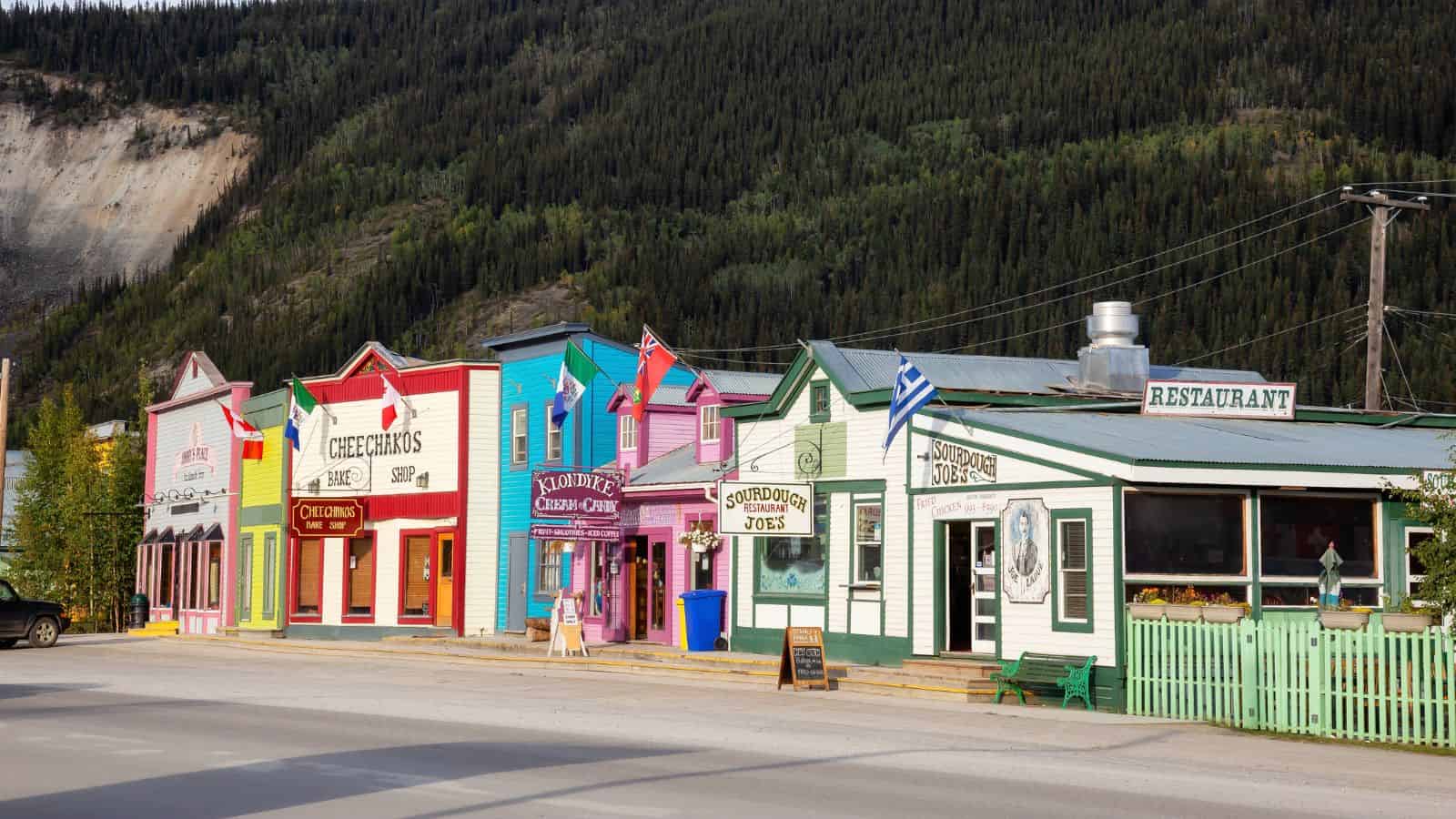 Colorful storefronts—a café, restaurant, and candy store—line a quiet street with forested hills behind them.