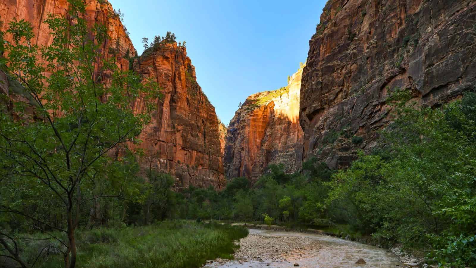 A scenic view of a canyon with tall, reddish rock cliffs on either side and a narrow river flowing through the middle. The sky is clear and blue, and the area is lush with green vegetation.