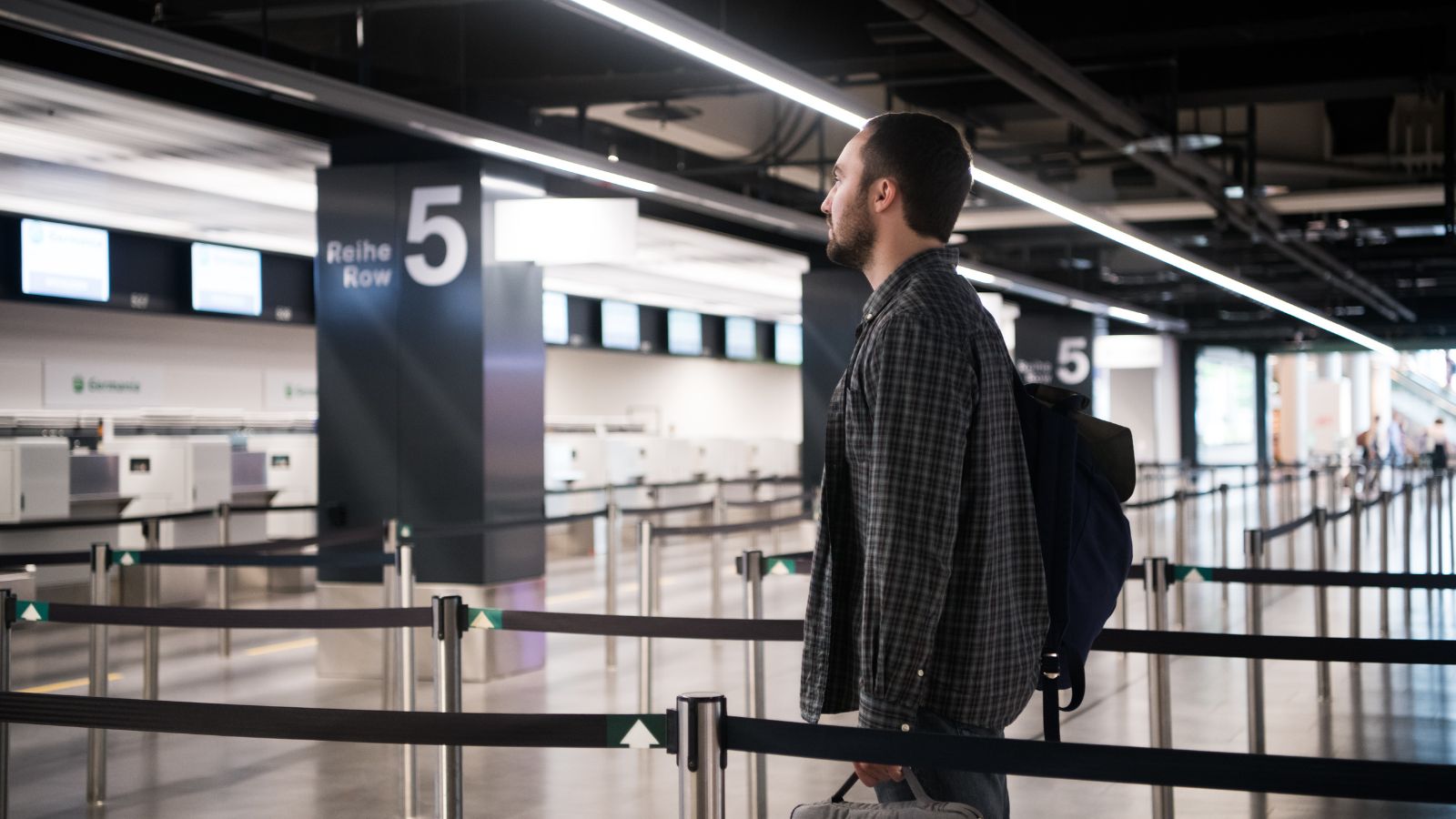A man with a backpack stands near row 5 in an empty airport check-in area, facing the counters.