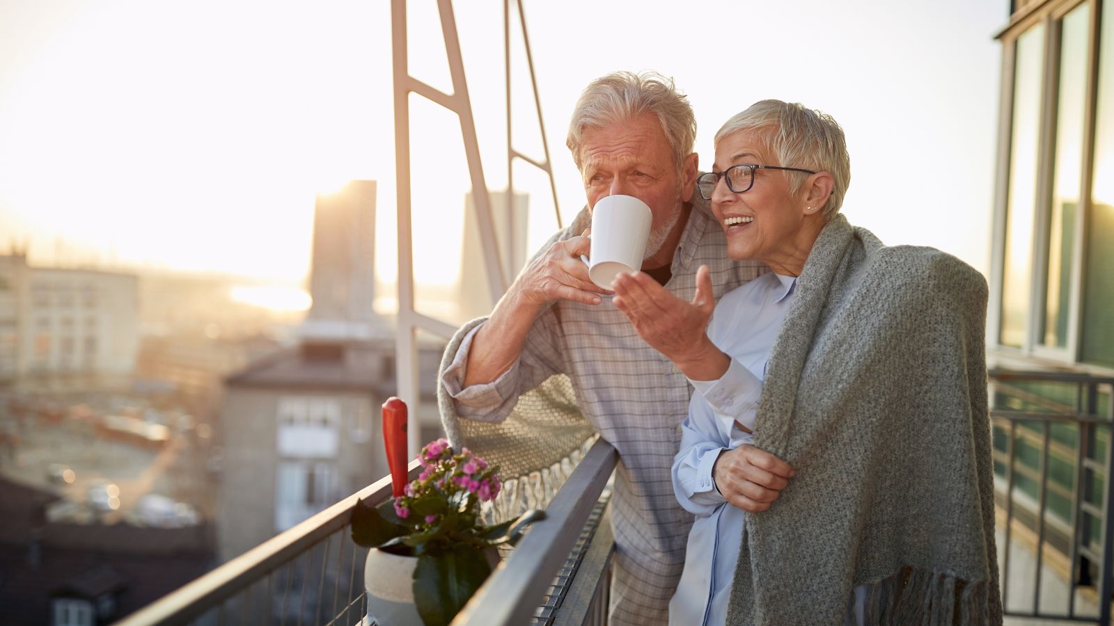 An older man sips from a mug on a balcony at sunset as a woman in a gray shawl smiles and gestures beside him.