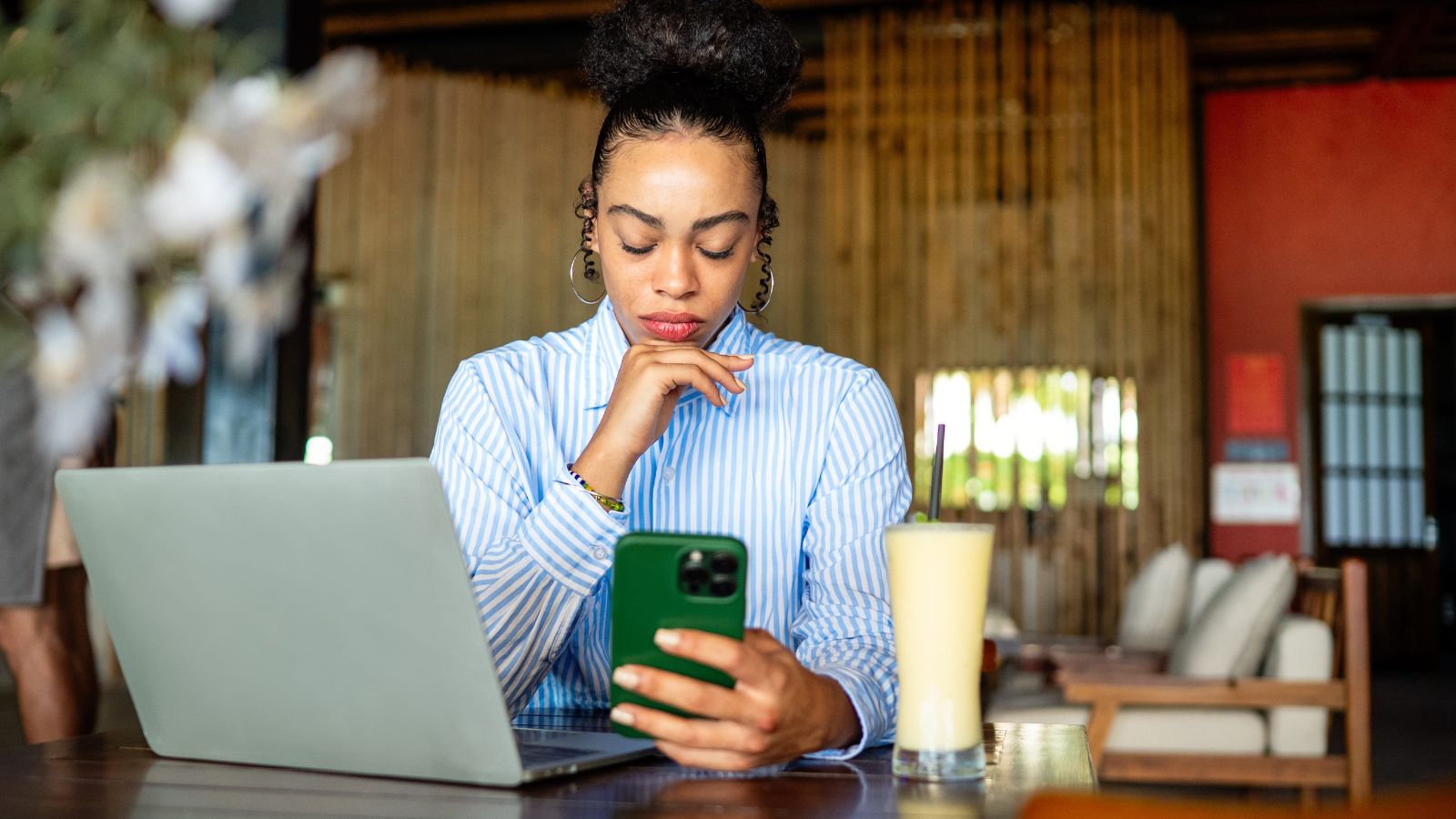 A photo of a woman, thinking deeply looking at her phone infront of her laptop.