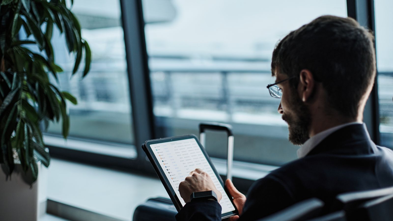 A man in business attire uses a tablet by an airport window, with a suitcase and potted plant nearby.