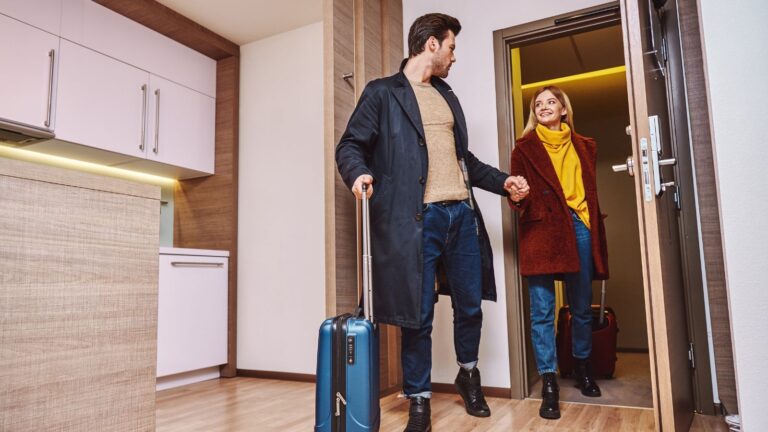 A man and woman in coats and casual clothes hold hands as they enter a modern apartment with luggage.