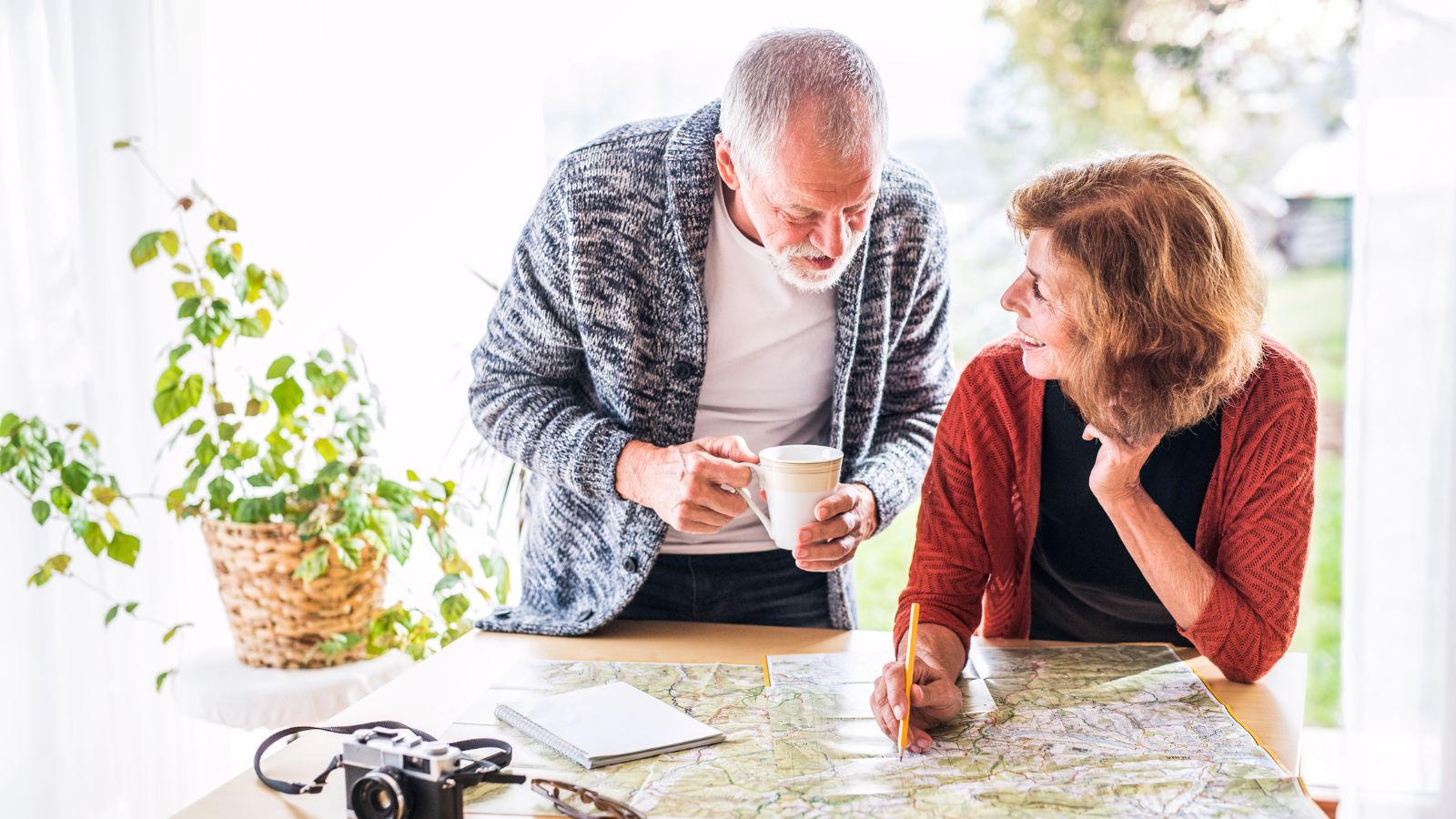 An older man and woman examine a map at a table with a mug, camera, notebook, plant, and the woman writes on the map.