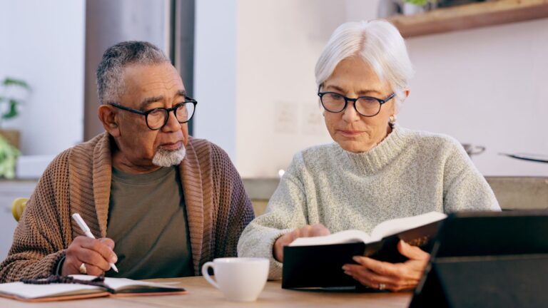 Two older adults with glasses sit at a table; one writes in a notebook, the other reads, with a coffee cup and tablet nearby.