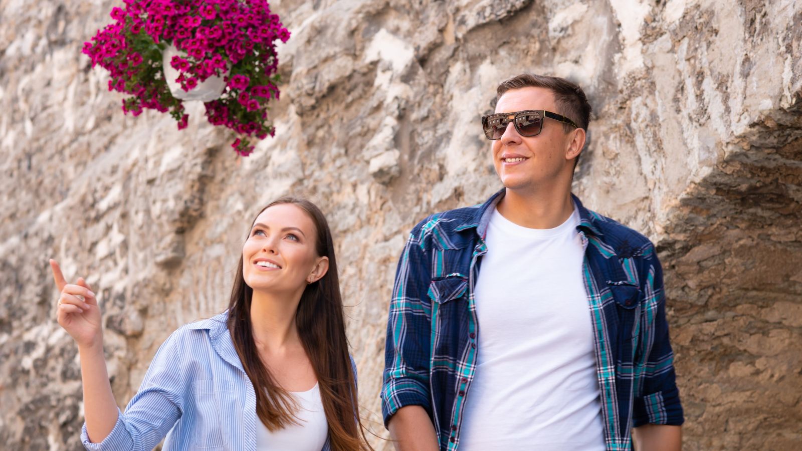 A woman and man walk by a stone wall, with the woman pointing at a hanging basket of purple flowers.