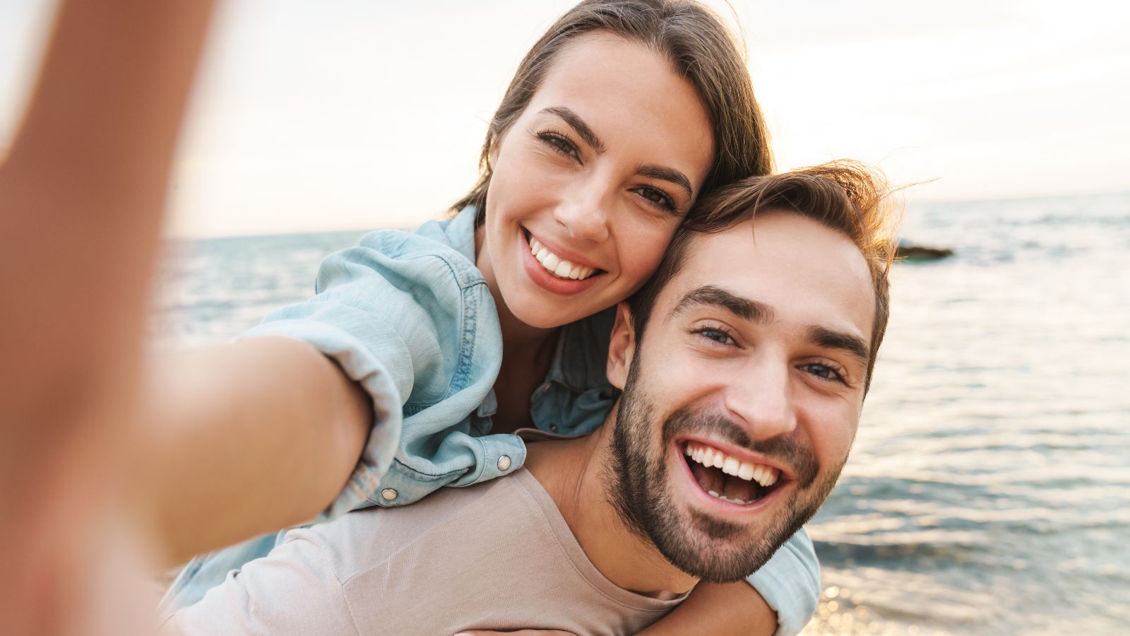 A smiling couple takes a beach selfie with the sea and sky in the background.