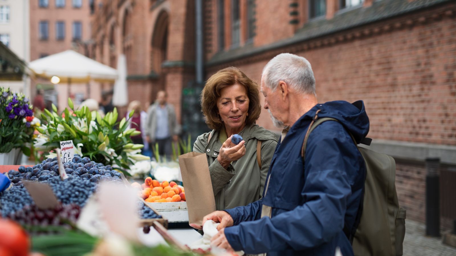 Two people examine plums and apricots at an outdoor market stall, with other shoppers and a brick building behind them.