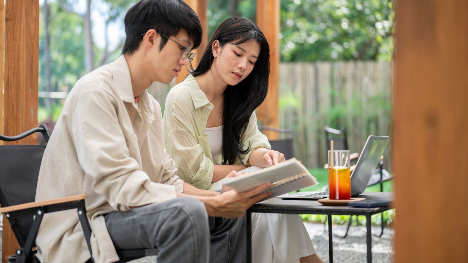 Two people sit at an outdoor table with a laptop, notebook, and drinks, appearing to review documents together.