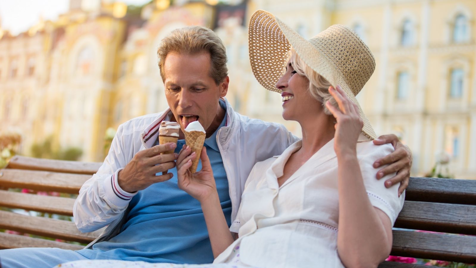 A smiling man and woman sit on an outdoor bench, sharing ice cream cones.