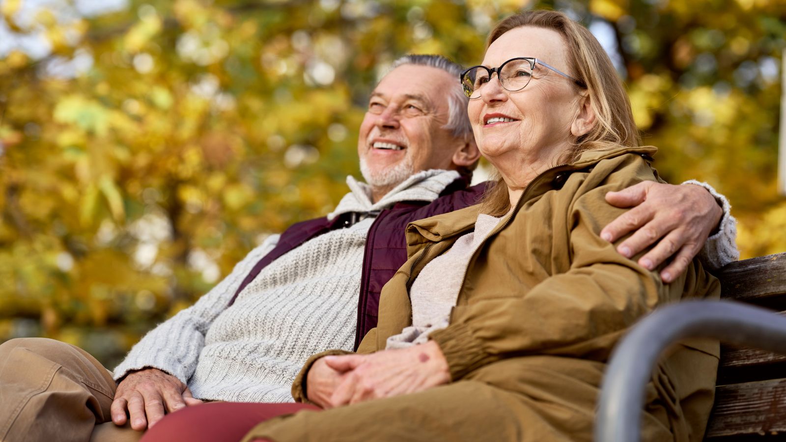 An older couple sits close on a bench outdoors, smiling amid autumn leaves.