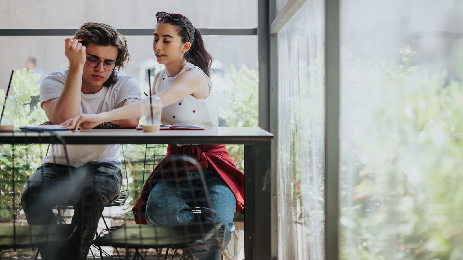 Two young adults sit by a window with drinks; one looks frustrated while the other points at something on the table, offering help.