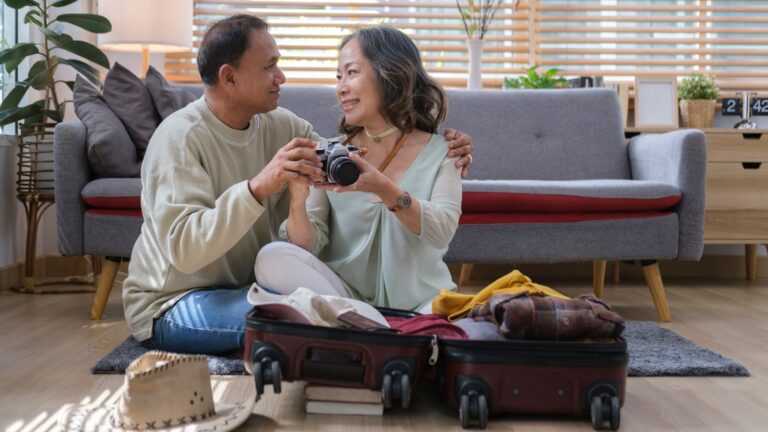 An older couple sits on the floor by an open suitcase, smiling at each other and holding a camera, with a sofa and plants nearby.