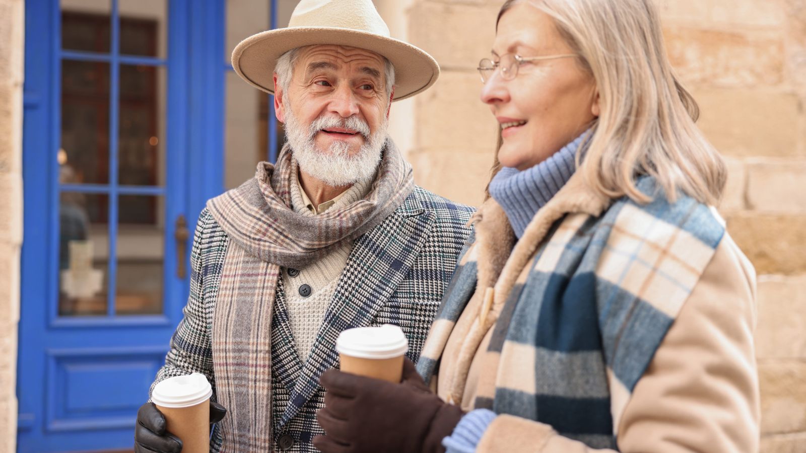 Two older adults in winter clothes stand outside by a blue-doored building, holding takeaway coffee and talking.
