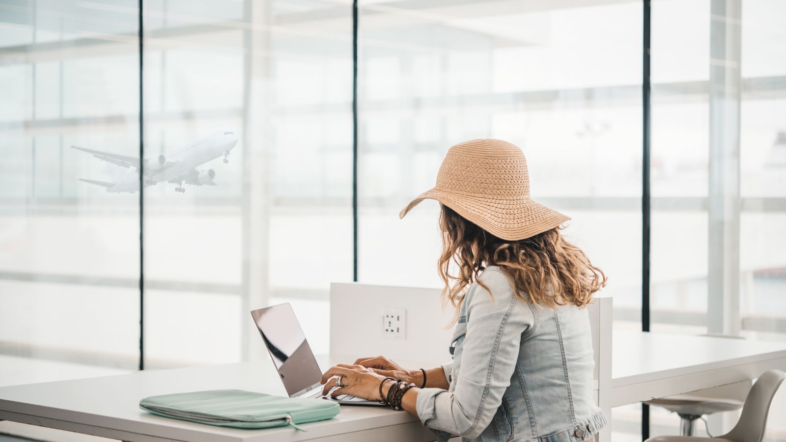 A woman in a sun hat uses a laptop at an airport terminal with a plane taking off outside large windows.