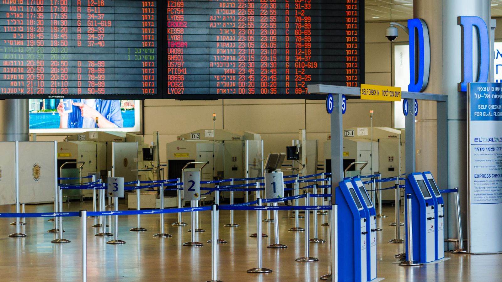 Empty airport check-in with self-service kiosks, queue barriers, and a large departures board showing flight information.