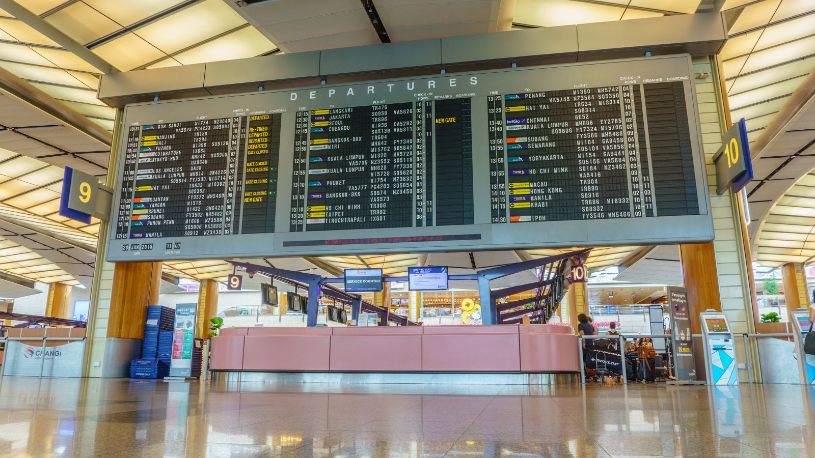 Electronic departures board shows flight info above check-in counters and passengers in an airport terminal.