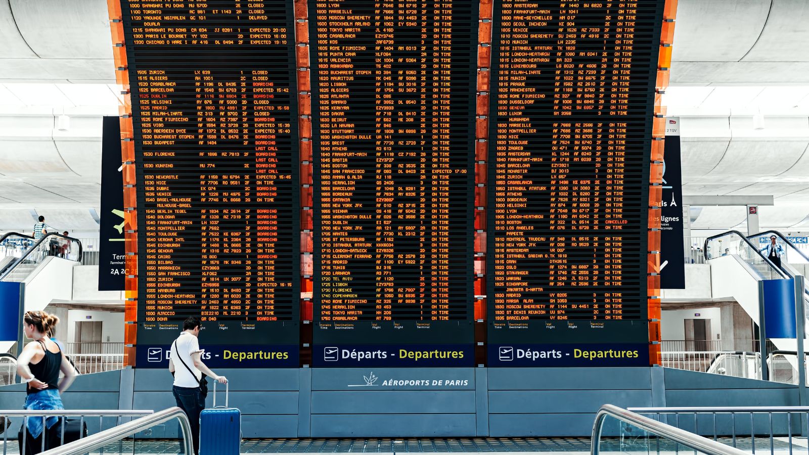 Travelers with luggage check a large digital airport departure board listing many flight times and destinations.