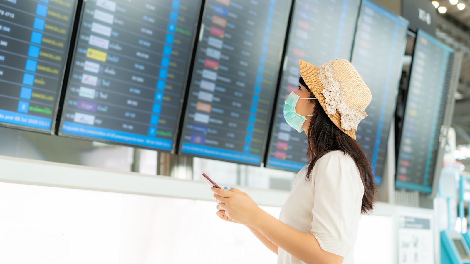 Woman in a hat and face mask checks her phone while viewing flight info displays at the airport.