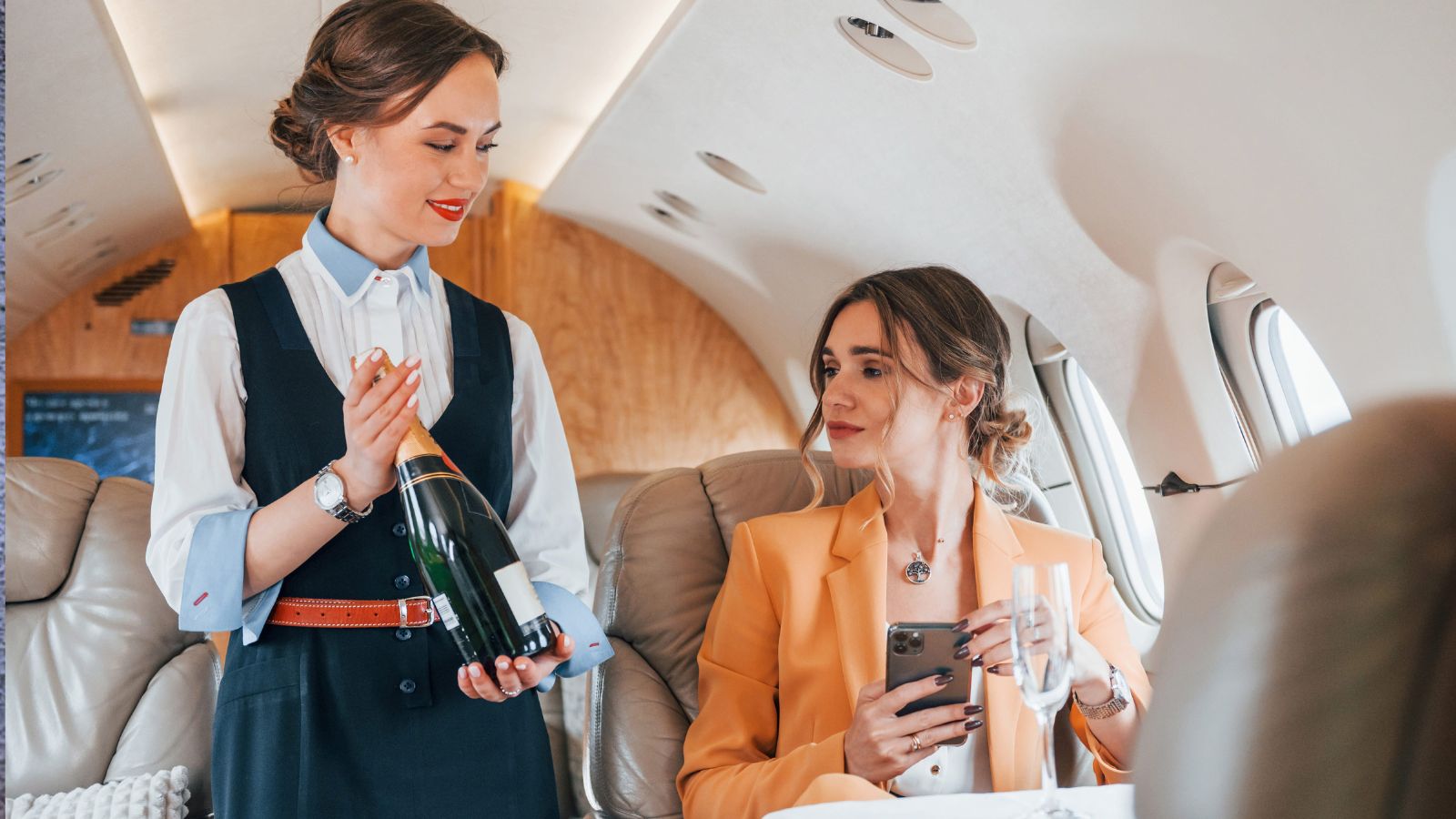 A formally dressed flight attendant offers champagne to a passenger holding a smartphone and glass on a private jet.