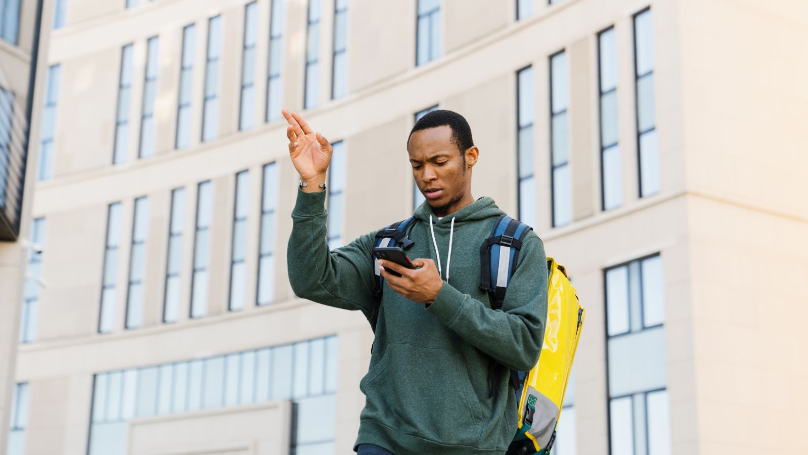 A man in a green hoodie and backpack stands outside a modern building, looking at his phone with one hand raised.