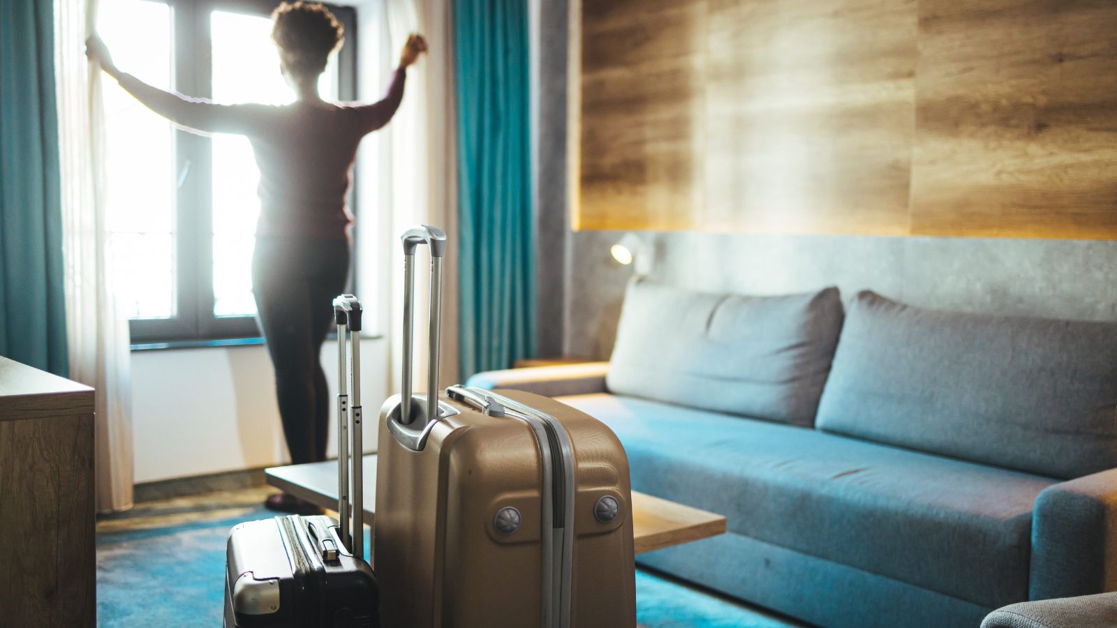 A person opens curtains by the window in a modern hotel room, with two suitcases near a grey sofa.