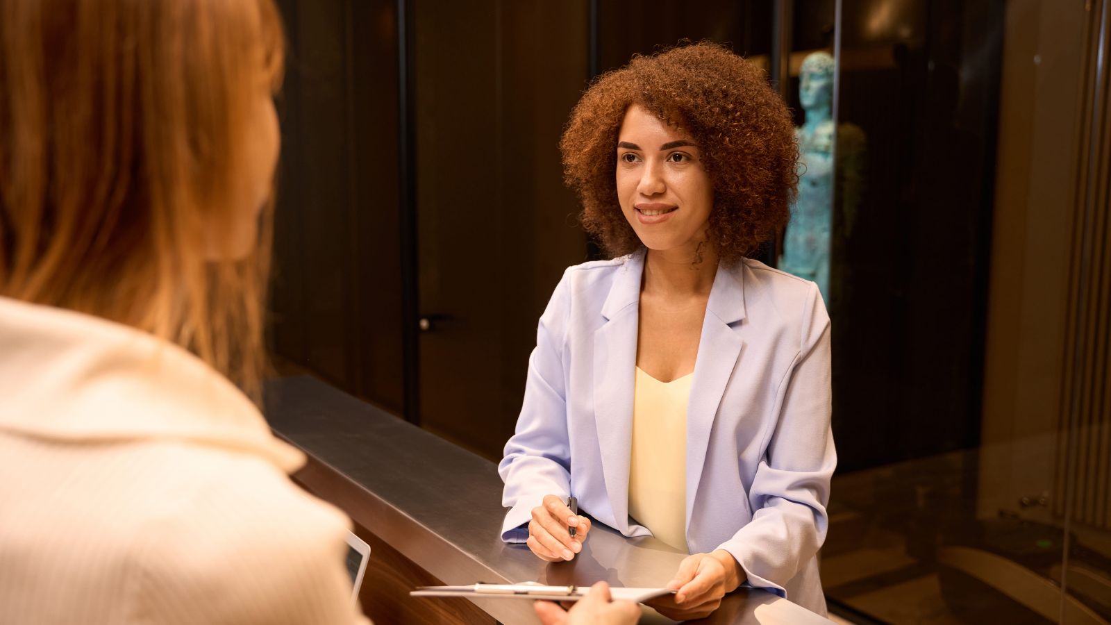 A photo of a “hotel guest checking in at front desk speaking with receptionist polite interaction modern hotel lobby candid.