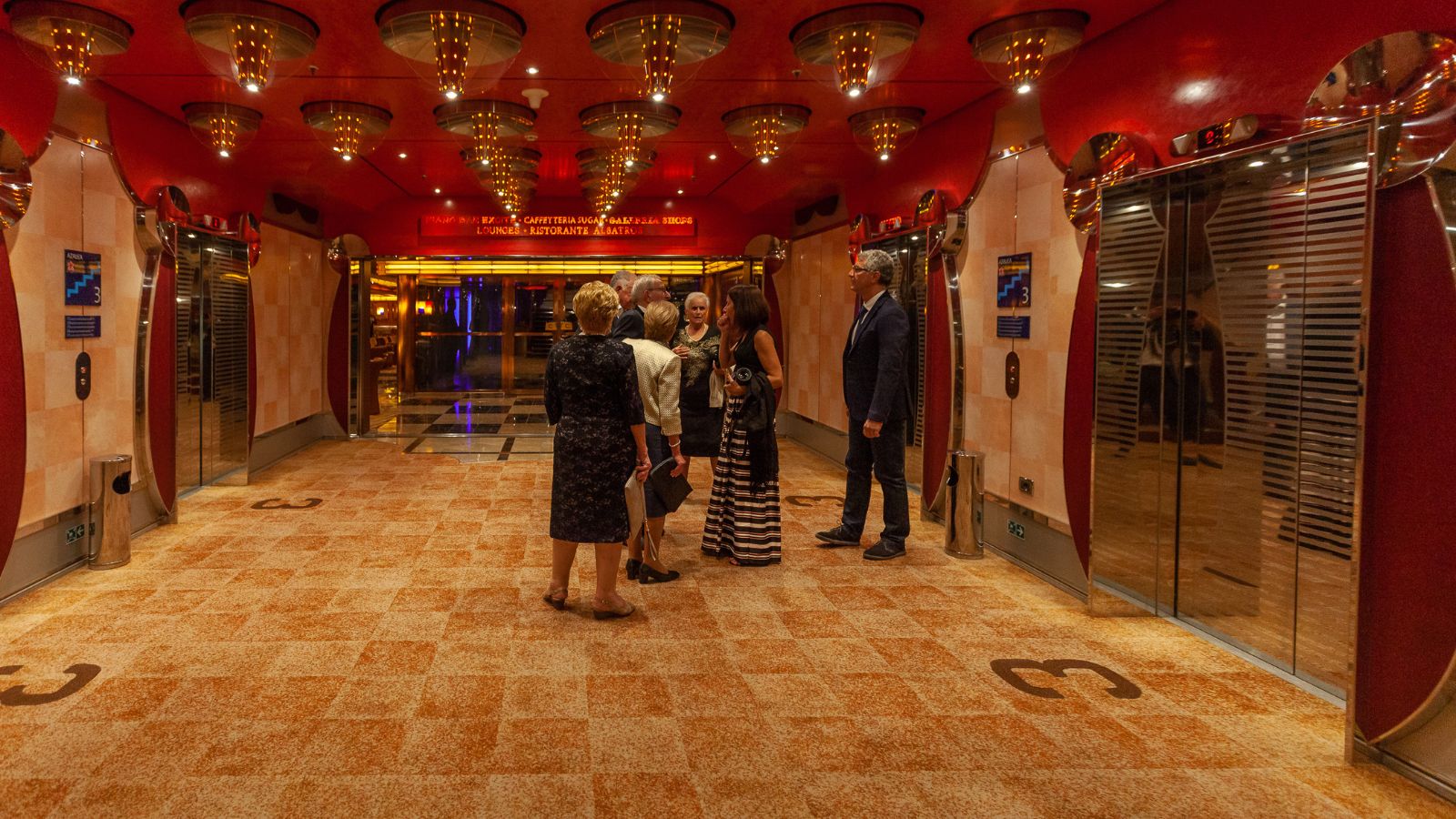 A photo cruise ship corridor near elevator bank with passengers walking toward theater entrance.