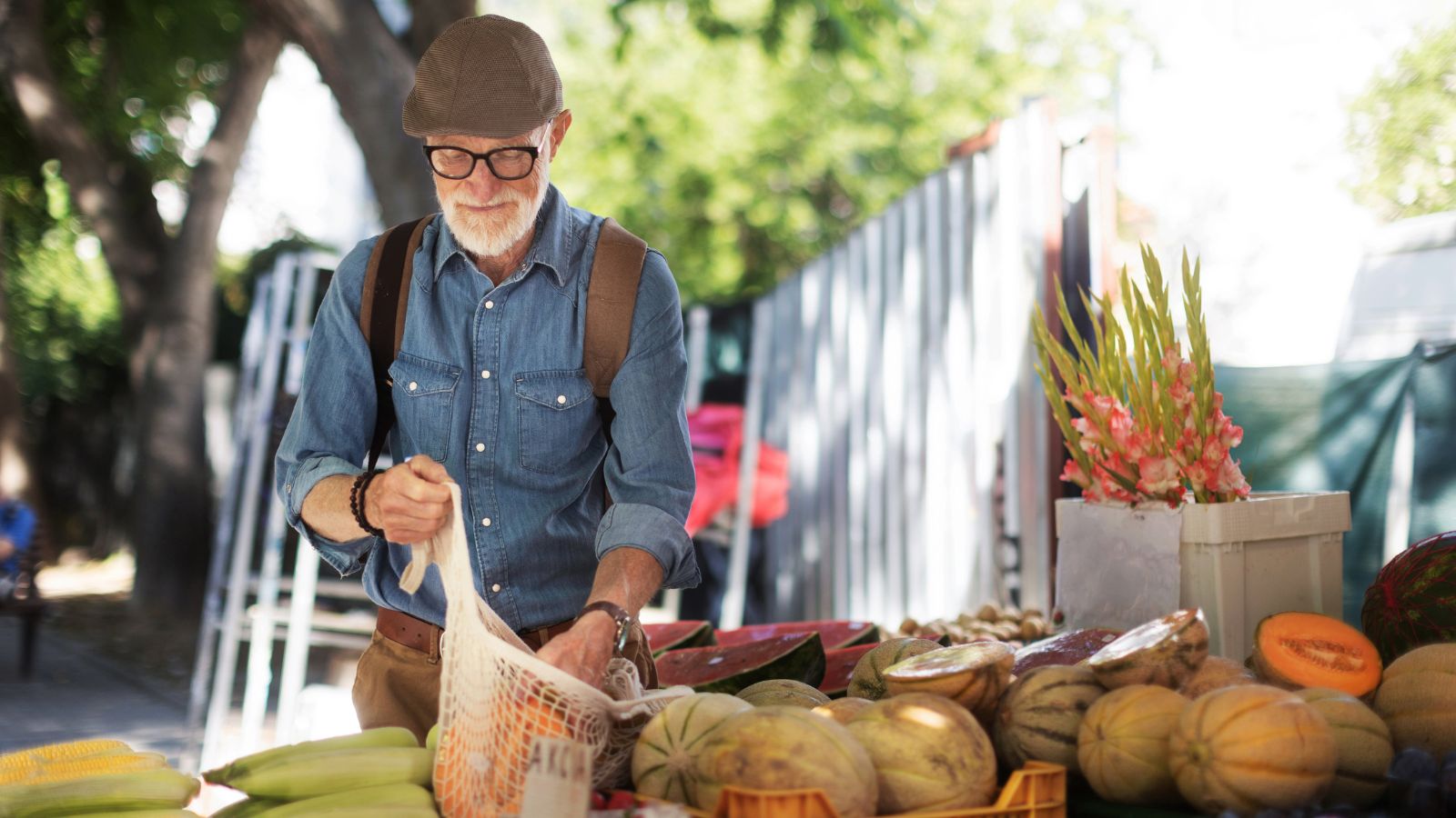 An older man wearing a cap and glasses buys fresh produce at an outdoor market, putting items into a reusable bag.