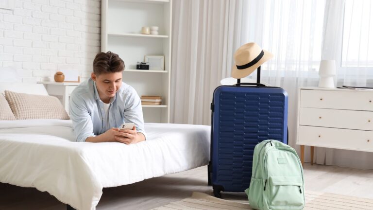 A photo of a man checking something in his phone, luggage behind hotel bed.