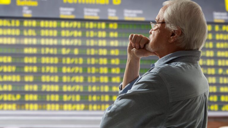 An older man in glasses gazes thoughtfully at a large airport departure board with yellow flight information.