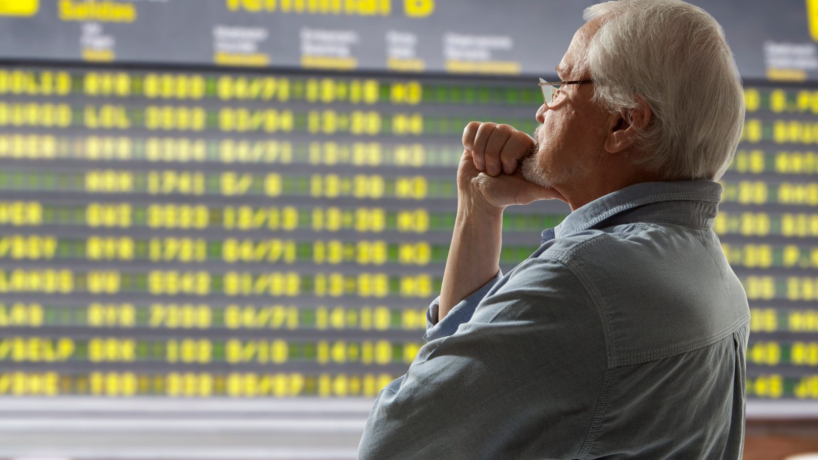 An older man in glasses gazes thoughtfully at a large airport departure board with yellow flight information.