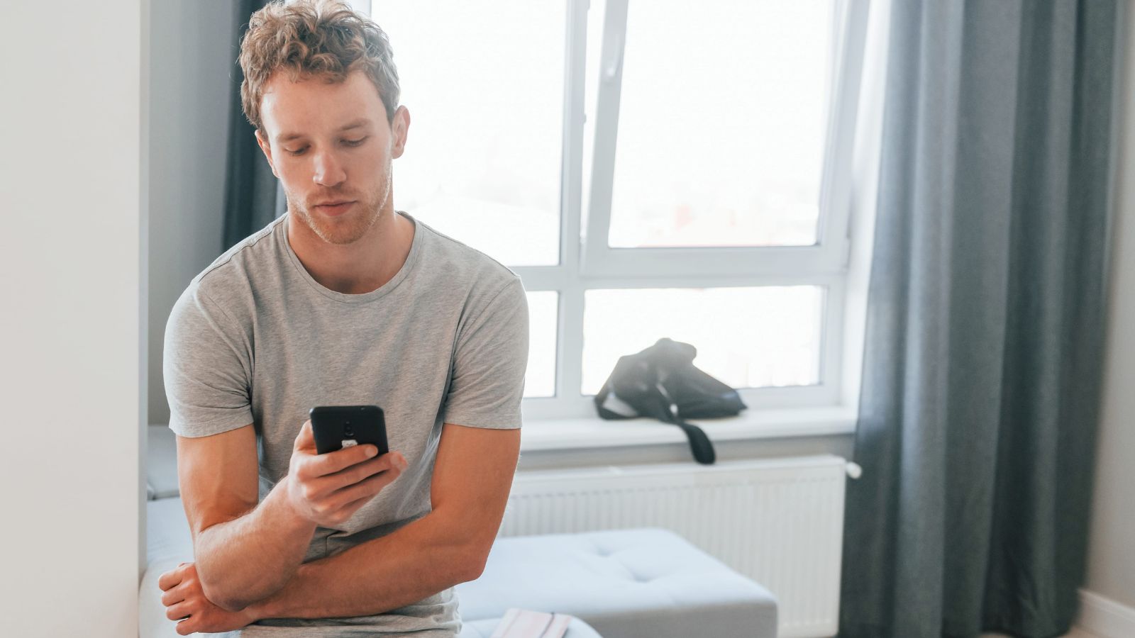 A photo of a traveler sitting on hotel bed in morning looking unsure checking phone without plan.
