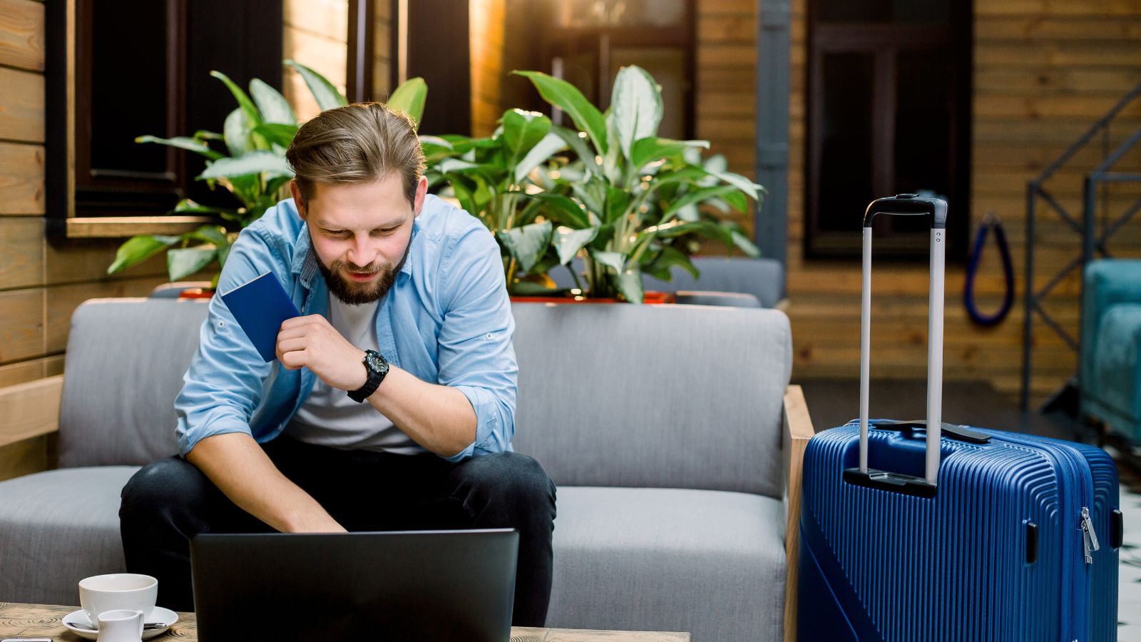 A man on a couch holds a passport and looks at his laptop, with a blue suitcase and cup on the nearby table.