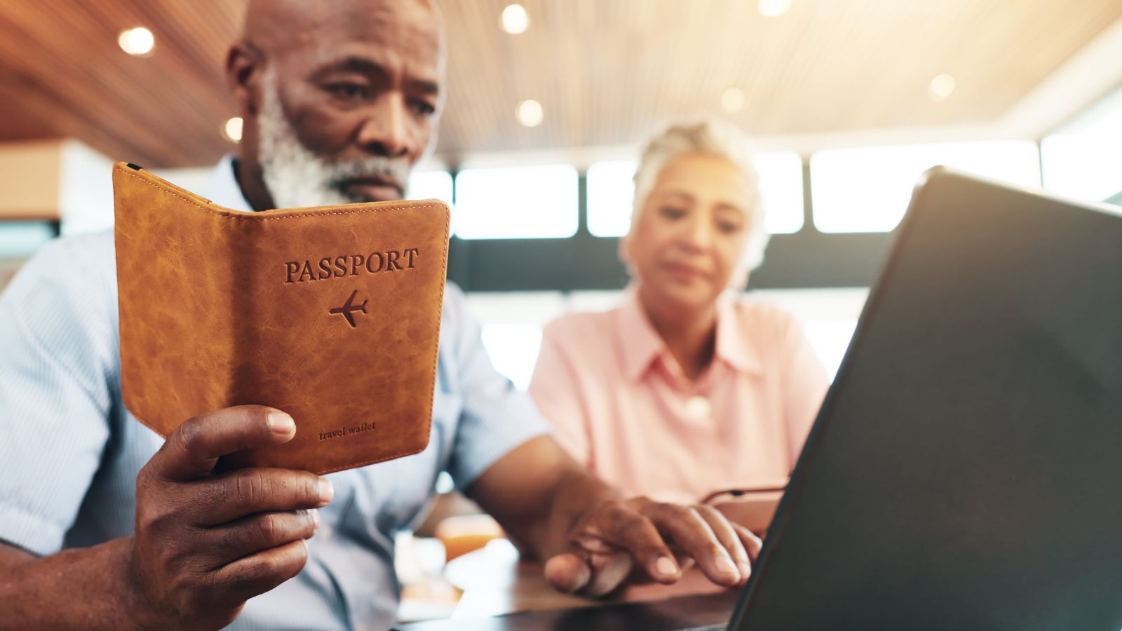 Two older adults at a table, one holding a passport, both looking at a laptop as they make travel plans.