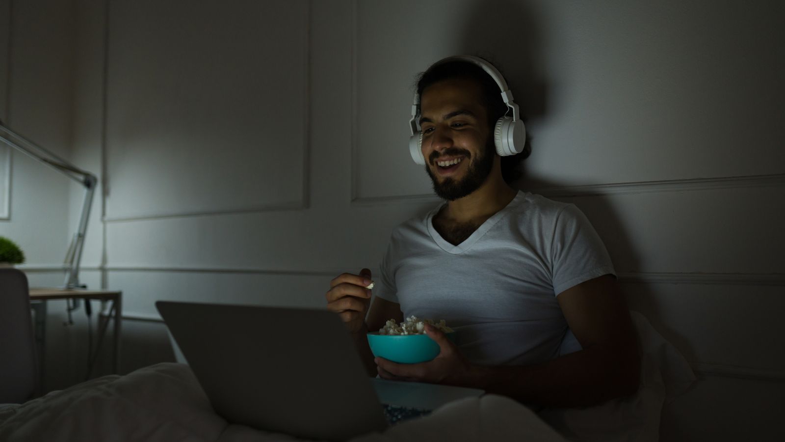 At night, a man in bed eats from a bowl and watches his laptop while wearing headphones.