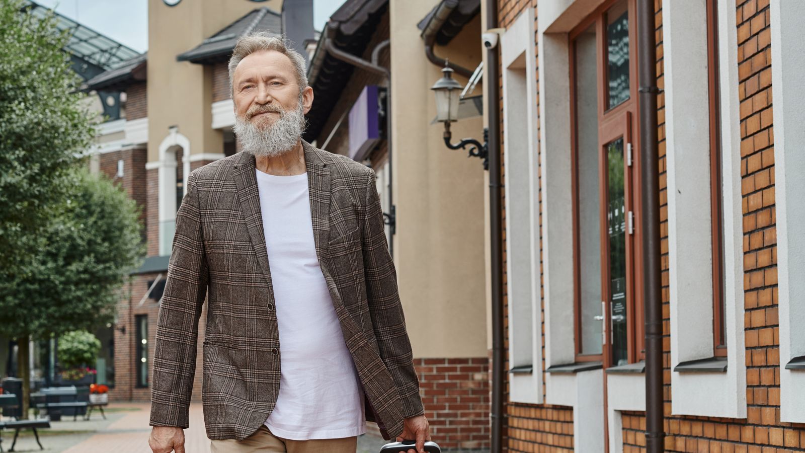 Older man with a gray beard in a checked blazer and white shirt walks along a street lined with buildings.