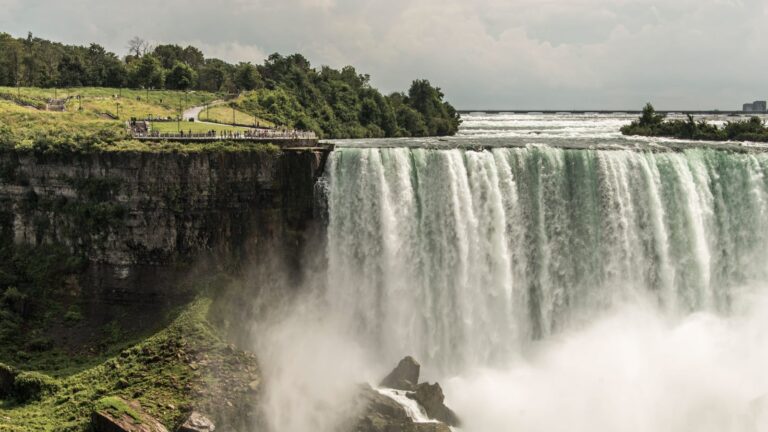 Water cascades over a rocky cliff surrounded by greenery as people stand on a nearby viewing platform.