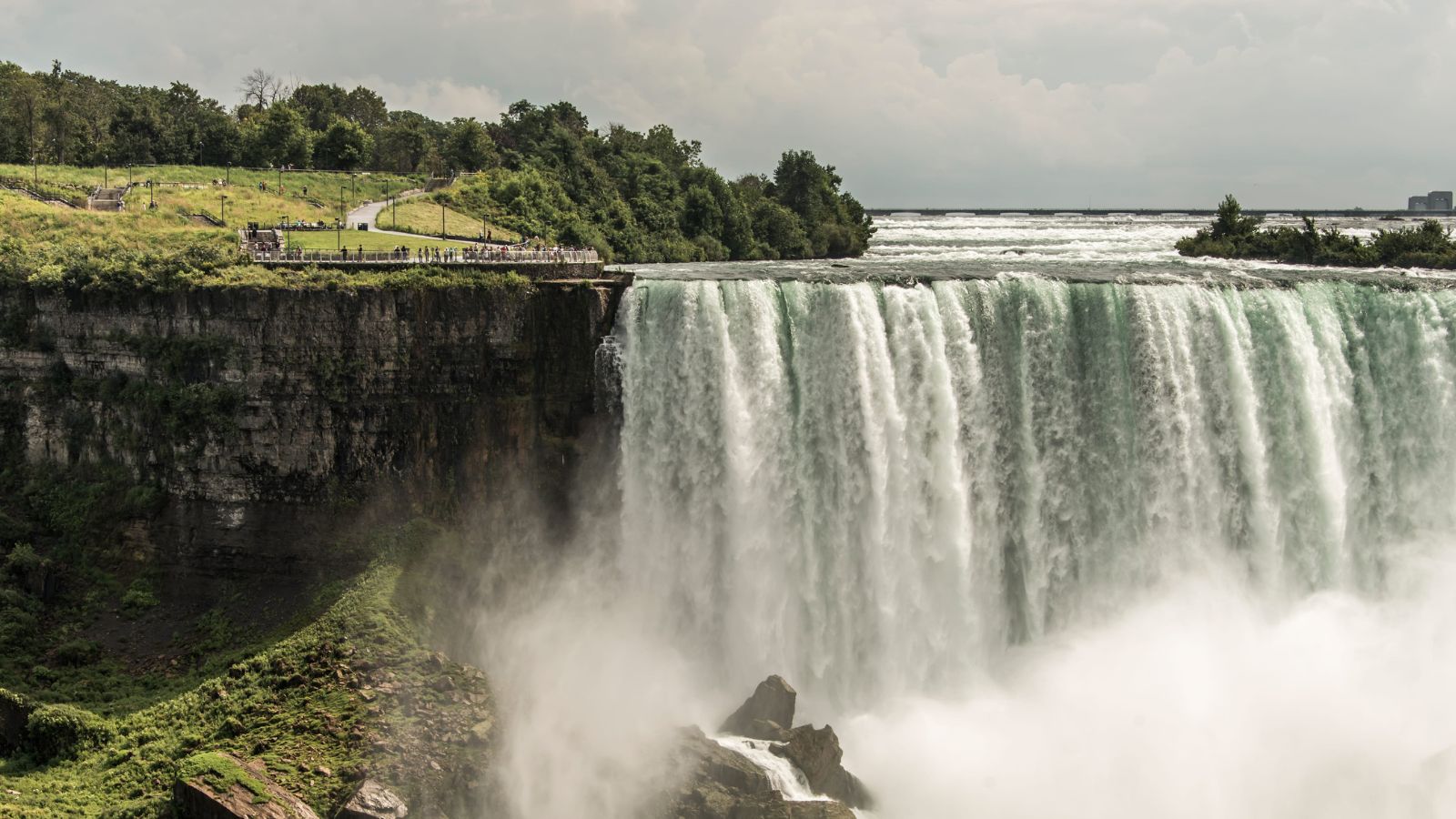 Water cascades over a rocky cliff surrounded by greenery as people stand on a nearby viewing platform.